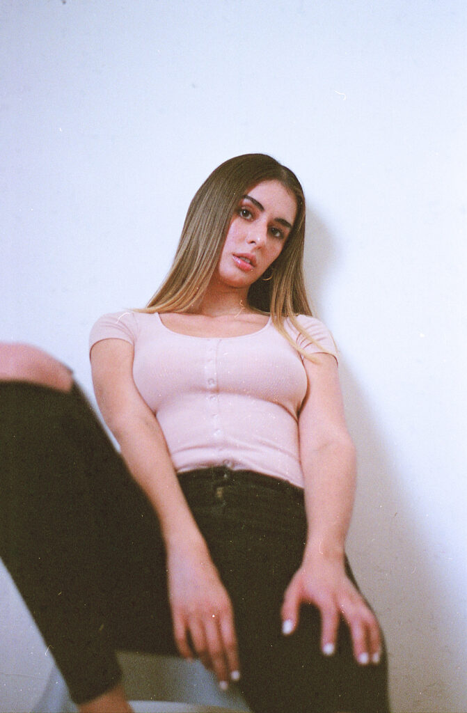 Film Photo of Marintia Bustos a model with long brown hair wearing a pink top and black jeans posed against a white wall.